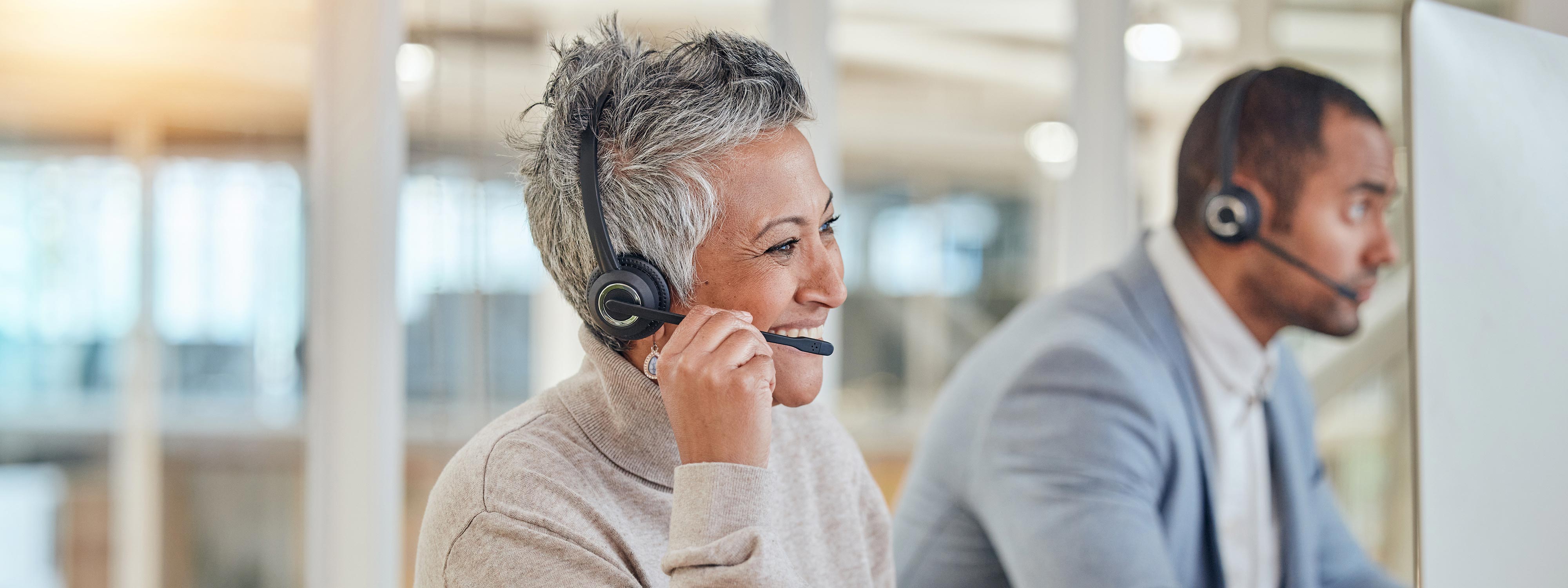 Das Bild zeigt zwei Callcenter-Mitarbeiter, die lächelnd und konzentriert miteinander interagieren und dabei Headsets tragen.

Im Vordergrund des Bildes ist eine Frau mittleren Alters mit kurzen, grauen Haaren zu sehen, die leicht zum Betrachter gedreht ist und in ihr Headset spricht. Sie trägt ein beiges Rollkragenoberteil und eine auffällige, ornamentale Ohrring. Ihr Gesichtsausdruck ist von einem freundlichen Lächeln und einer offensichtlichen Freude an ihrer Tätigkeit geprägt, ihre Augen sind leicht zusammengekniffen, was auf eine positive Interaktion hindeutet. Ihre linke Hand nähert sich dem Mikrofon ihres Headsets, was darauf hindeutet, dass sie gerade spricht oder eine Unterbrechung erwägt.

Im Hintergrund und leicht unscharf befindet sich ein Mann, der ebenfalls ein Headset trägt und seinem Gegenüber aufmerksam zuzuhören scheint. Er ist mit einem hellblauen Sakko über einem weißen Hemd bekleidet und blickt nach rechts, möglicherweise auf einen Bildschirm oder zu seinem Gegenüber. Sein Gesichtsausdruck ist konzentriert und professionell.

Die Atmosphäre des Bildes ist hell und professionell, mit weichem Licht, das von links einfällt und eine warme Ausleuchtung schafft. Der Hintergrund ist unscharf, aber es scheint sich um ein modernes Büroumfeld mit Fenstern oder Glasabtrennungen zu handeln. Die Stimmung ist positiv und produktiv, was die Zufriedenheit und das Engagement der dargestellten Personen vermittelt. Es suggeriert ein positives Arbeitsumfeld, in dem Kommunikation und Kundenservice im Vordergrund stehen.