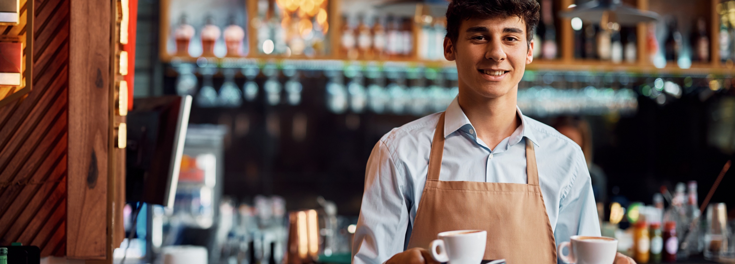 Ein junger Mann in einer Schürze lächelt in die Kamera und hält zwei Tassen Cappuccino in der Hand, während er in einem Café steht.

Das Bild zeigt einen jungen Mann, vermutlich einen Barista, der frontal und leicht von der Seite lächelnd in die Kamera blickt. Er hat dunkle, kurz geschnittene Haare und trägt ein hellblaues Hemd unter einer braunen Schürze. Seine Hände halten eine kleine schwarze Untertasse mit zwei weißen Cappuccinotassen darauf. Das Getränk in den Tassen ist hellbraun mit weißem Milchschaum.

Der Hintergrund ist unscharf, aber es ist erkennbar, dass er sich in einem Café oder einer Bar befindet. Hinter dem Mann sind Regale mit Flaschen, Gläsern und anderen Barutensilien zu sehen. Die Beleuchtung im Hintergrund ist warm und diffus, mit einigen Lichtern, die als Bokeh-Effekte erscheinen. Links im Bild ist ein Teil einer hölzernen Theke oder eines dekorativen Elements mit schrägen Linien zu erkennen.

Die Atmosphäre ist freundlich und einladend. Das Lächeln des Mannes und die präsentierte Kaffeespezialität vermitteln ein Gefühl von Service und Genuss. Die warme Beleuchtung trägt zur Gemütlichkeit bei und deutet auf eine angenehme Stimmung hin, die man in einem Café erwarten würde. Es ist ein Bild, das Gastfreundschaft und die Freude am Kaffee vermittelt.