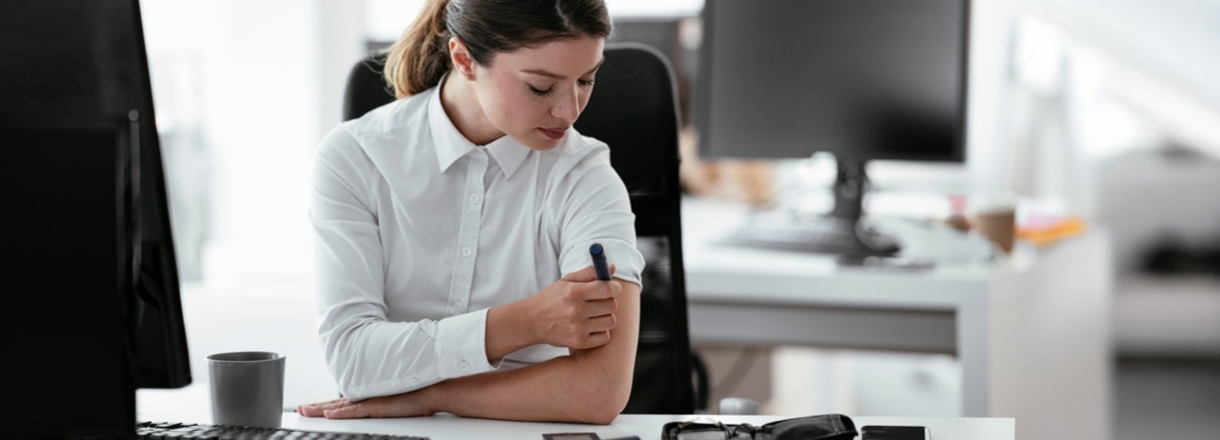 Eine junge Frau im Büro injiziert sich Insulin, während sie auf ihren Computerbildschirm blickt.

Das Bild zeigt eine junge Frau, die in einem modernen Büro sitzt. Sie trägt ein weißes Hemd mit aufgekrempelten Ärmeln und ihre Haare sind zu einem Pferdeschwanz zusammengebunden. Ihr Blick ist nach unten auf ihren linken Oberarm gerichtet, wo sie sich mit einem blauen Injektionsstift Insulin spritzt. Ihre Gesichtsausdruck ist konzentriert, aber gefasst, was auf eine routinemäßige Handlung hindeutet. Sie sitzt an einem Schreibtisch, auf dem ein Computerbildschirm, eine Tastatur und eine graue Tasse zu sehen sind. Im Hintergrund sind weitere Büromöbel und Computerbildschirme angedeutet, die eine professionelle Arbeitsumgebung darstellen. Die Beleuchtung ist hell und diffus, was eine klare und sachliche Atmosphäre schafft, die jedoch durch die persönliche Handlung der Frau eine tiefere Ebene erhält. Die Stimmung ist ruhig und unaufgeregt, was die Normalität des Lebens mit einer chronischen Krankheit im Arbeitsalltag unterstreicht. Das Bild thematisiert die Herausforderungen und die Selbstständigkeit von Menschen, die trotz gesundheitlicher Einschränkungen ein aktives Berufsleben führen.