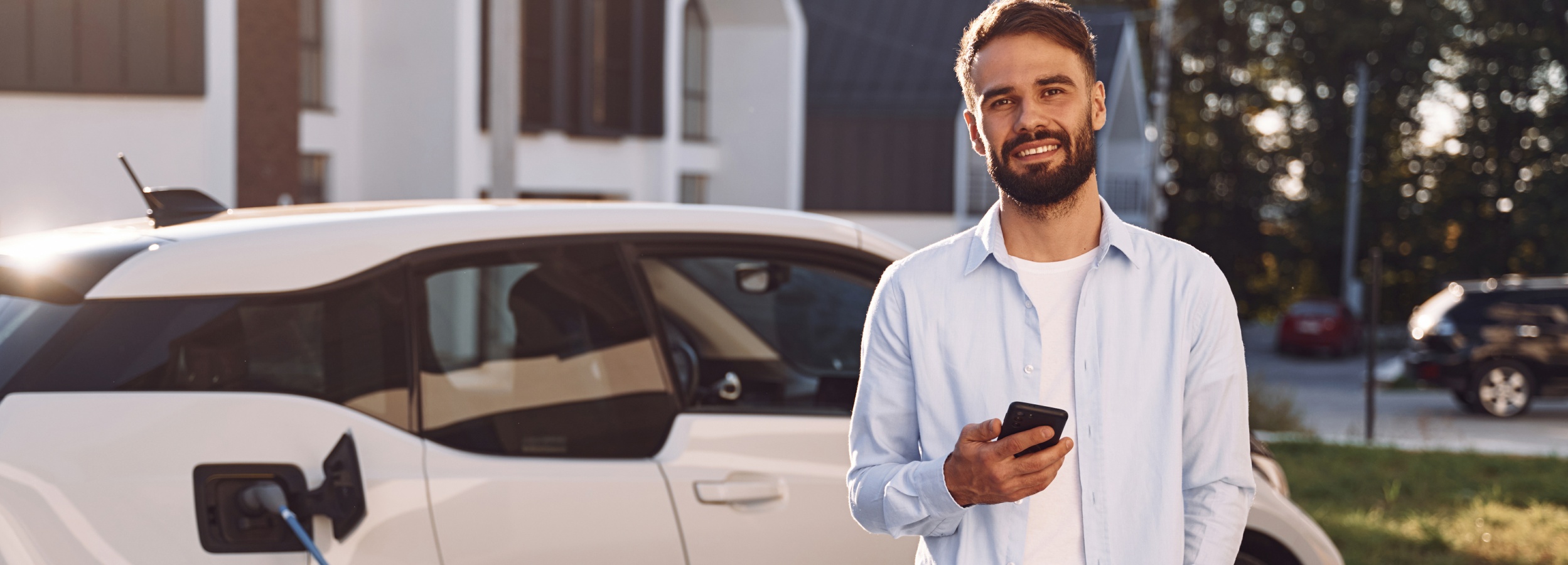 Ein lächelnder Mann hält sein Smartphone in der Hand, während sein Elektroauto im Hintergrund geladen wird.

Auf dem Bild ist ein junger Mann mit einem Bart zu sehen, der ein hellblaues Hemd über einem weißen T-Shirt trägt. Er steht im Freien vor einem modernen Gebäude und hält lächelnd ein Smartphone in seiner rechten Hand. Seine linke Hand ruht locker auf seiner Hüfte. Im Vordergrund befindet sich ein weißes Elektroauto, dessen Ladeanschluss geöffnet ist und ein blaues Ladekabel eingesteckt hat. Die Sonne scheint von rechts und wirft ein warmes Licht auf den Mann und das Auto. Im Hintergrund sind weitere Häuser und Bäume zu erkennen, sowie ein dunkles Auto auf der Straße. Die Atmosphäre ist friedlich und positiv, was durch das Lächeln des Mannes und die warme Beleuchtung unterstrichen wird. Das Bild vermittelt den Eindruck von Nachhaltigkeit und moderner Technologie, die nahtlos in den Alltag integriert ist.