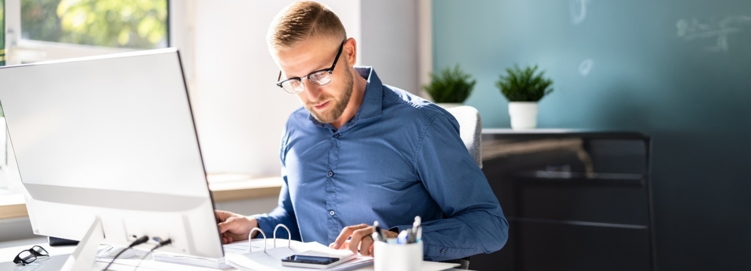 Ein Mann in einem blauen Hemd sitzt an seinem Schreibtisch vor einem Computer und studiert Dokumente.

Auf dem Bild ist ein Mann mit kurzen blonden Haaren und einem Dreitagebart zu sehen, der eine Brille mit schwarzem Rahmen trägt und konzentriert auf Papiere starrt, die vor ihm auf einem Schreibtisch liegen. Er trägt ein blaues Hemd mit kleinen Punkten. Seine rechte Hand ruht auf den Dokumenten, während seine linke auf dem Schreibtisch liegt. Vor ihm steht ein großer, weißer Computermonitor. Links neben dem Monitor liegt eine Brille. Auf dem Schreibtisch befinden sich außerdem ein Smartphone, ein weißer Becher mit Schreibutensilien und ein Ordner. Im Hintergrund sind zwei kleine Topfpflanzen auf einem Regal zu erkennen, sowie eine dunkelblaue Wand, an der schemenhaft etwas geschrieben steht. Durch ein Fenster fällt helles Tageslicht herein, das einen Teil des Raumes beleuchtet und eine helle, konzentrierte Atmosphäre schafft. Die Stimmung ist ruhig und fokussiert, was auf eine ernsthafte Arbeitsatmosphäre hindeutet.