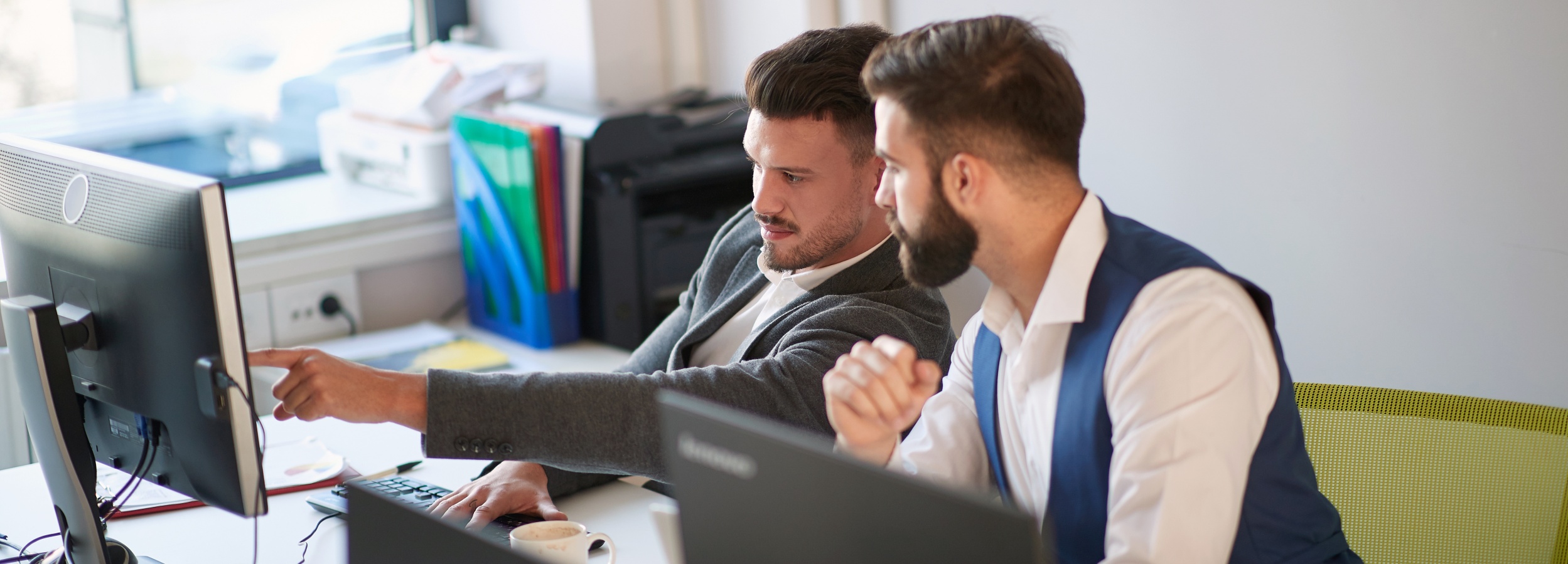 Zwei konzentrierte Männer, die in einem Büro an einem Computer arbeiten und ihn diskutieren.

Das Bild zeigt zwei Männer in einem Büro, die sich über einen Computermonitor beugen. Der Mann auf der linken Seite trägt einen grauen Anzug und ein weißes Hemd und zeigt mit dem Finger auf den Bildschirm, während er spricht. Er hat dunkles Haar, das auf die Seite gekämmt ist, und einen gepflegten Bart. Der Mann auf der rechten Seite trägt eine dunkelblaue Weste über einem weißen Hemd und hat einen braunen Bart und dunkles Haar. Beide Männer scheinen in das, was sie auf dem Bildschirm sehen, vertieft zu sein.

Der Schreibtisch, an dem sie sitzen, ist ordentlich, mit einem weiteren Computermonitor, einer Tastatur, einer Maus und einer Kaffeetasse. Im Hintergrund sind Ordner in leuchtenden Farben, ein Drucker und ein Fenster zu sehen, das Tageslicht hereinlässt. Die Atmosphäre ist professionell und konzentriert, mit einem Hauch von Kollaboration. Die Beleuchtung ist hell und natürlich, was zu einer sauberen und modernen Büroumgebung beiträgt. Die Gesamtstimmung ist eine der Arbeitseffizienz und des Teamworks.