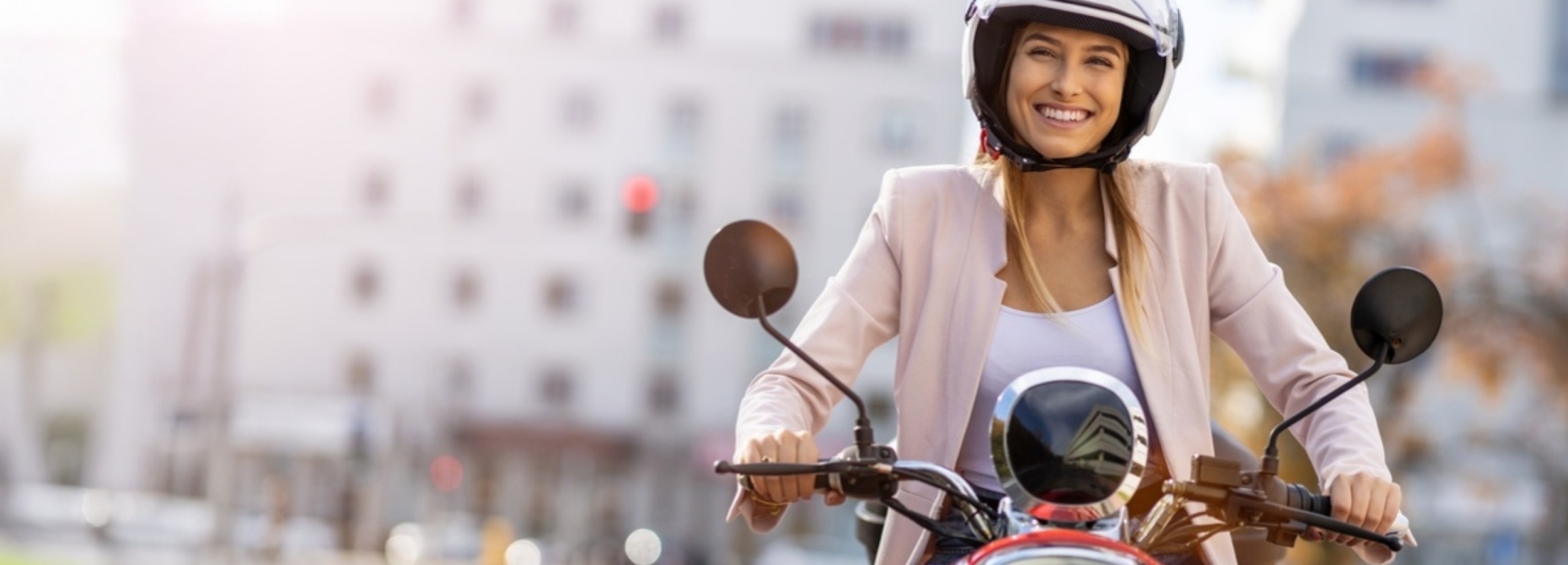 Eine junge Frau fährt lächelnd auf einem roten Roller durch die Stadt.

Das Bild zeigt eine junge Frau, die einen weißen Motorradhelm trägt und auf einem auffällig roten Roller sitzt. Sie hat blonde Haare, die unter dem Helm hervorfallen, und trägt eine helle, rosafarbene Jacke über einem weißen Top. Ihr Gesichtsausdruck ist strahlend und freudig, mit einem breiten Lächeln, das ihre Begeisterung für die Fahrt zeigt. Ihre Hände halten den Lenker des Rollers, wobei ihre linke ihren Daumen auf dem Gasgriff ruht. Der Roller ist detailreich dargestellt, mit Chromakzenten und einem runden Scheinwerfer, der leicht nach oben geneigt ist. Im Spiegel des Rollers spiegelt sich ein Gebäude.

Die Umgebung ist unscharf und deutet auf eine städtische Szenerie hin. Im Hintergrund sind unscharfe Silhouetten von Gebäuden zu sehen, einige mit dunklen Fenstern. Ein roter Lichtpunkt, möglicherweise eine Ampel oder ein Bremslicht, ist im Hintergrund erkennbar. Die Lichtverhältnisse suggerieren einen sonnigen Tag, mit weichem, natürlichem Licht, das die Szene erhellt und eine warme, fröhliche Atmosphäre schafft. Die Stimmung ist unbeschwert und dynamisch, vermittelt ein Gefühl von Freiheit und Freude am Unterwegssein in der Stadt.