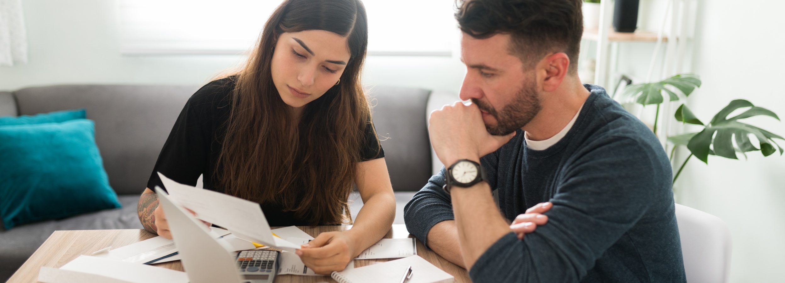 Zwei junge Menschen sitzen an einem Tisch und besprechen Unterlagen, wobei eine ruhige und etwas angespannte Atmosphäre herrscht.

Das Bild zeigt zwei Personen, eine junge Frau und einen jungen Mann, die an einem Holztisch sitzen und sich offensichtlich mit Finanzunterlagen beschäftigen. Die Frau, mit langen, dunkelbraunen Haaren, trägt ein schwarzes T-Shirt und schaut konzentriert auf ein Blatt Papier in ihren Händen, das sie mit beiden Händen hält. Ihre Miene ist ernst, und ihr Blick ist nach unten gerichtet, was auf tiefe Konzentration oder Besorgnis hindeutet. Auf ihrem linken Unterarm sind Tätowierungen sichtbar. Neben den Papieren liegt ein Taschenrechner, und ein Notizbuch mit einem Stift ist ebenfalls auf dem Tisch zu sehen.

Der Mann, mit kurz geschnittenem dunklem Haar und Bart, trägt einen dunkelblauen Pullover über einem weißen T-Shirt. Er sitzt mit dem Oberkörper leicht nach vorne gebeugt und hat seine rechte Hand nachdenklich an sein Kinn gelegt. Sein Blick ist ebenfalls auf die Unterlagen gerichtet, aber seine Augen scheinen leicht glasig, und seine Stirn ist leicht gerunzelt, was auf Nachdenklichkeit oder auch Frustration hindeuten könnte. Er trägt eine Armbanduhr mit weißem Zifferblatt und schwarzem Armband am linken Handgelenk.

Der Hintergrund des Bildes ist hell und aufgeräumt. Im linken Teil des Bildes ist ein Teil eines grauen Sofas mit zwei auffälligen, türkisfarbenen Kissen zu erkennen. Im rechten Hintergrund steht ein weißes Regal, auf dem in einem schwarzen Topf eine Zimmerpflanze mit großen, geschlitzten Blättern steht. Die Beleuchtung im Raum ist weich und natürlich, was zu einer eher intimen, aber auch ernsten Atmosphäre beiträgt.

Die Stimmung des Bildes ist geprägt von Ernsthaftigkeit und Konzentration. Es scheint, als ob die beiden Personen eine wichtige finanzielle Angelegenheit besprechen, sei es die Verwaltung von Haushaltsgeldern, die Planung von Ausgaben oder die Lösung eines finanziellen Problems. Die Körpersprache und die Mimik beider Personen deuten auf eine gewisse Anspannung oder Sorge hin, aber auch auf den gemeinsamen Willen, die Situation zu bewältigen. Das Bild fängt einen Moment der gemeinsamen Verantwortung und des ernsten Gesprächs ein.