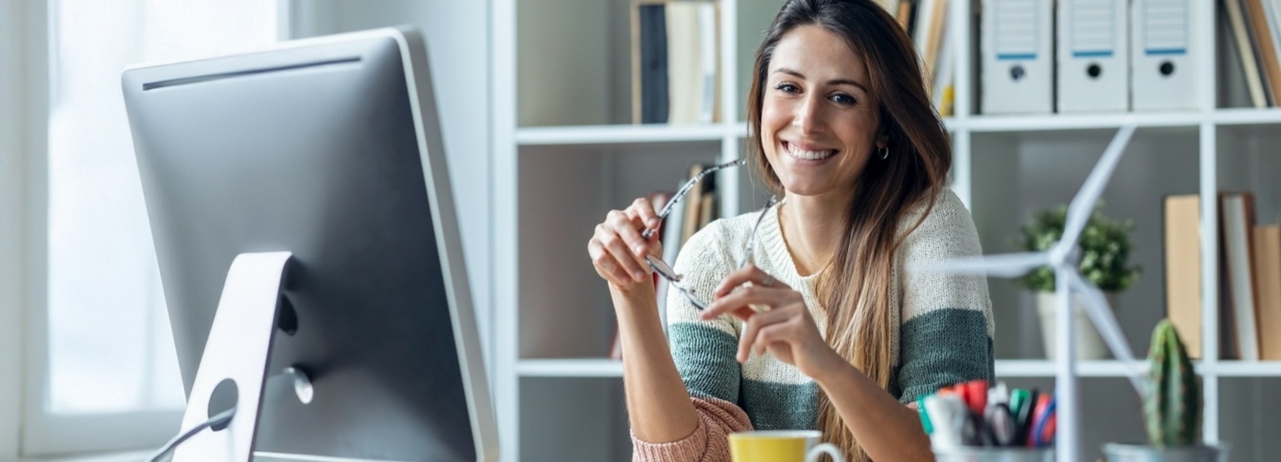 Eine lächelnde junge Frau mit Brille in einem hellen Büro mit einem Computer und einem Regal voller Büromaterialien.

Das Bild zeigt eine junge Frau, die in einem modernen Büro sitzt und von der Seite lächelnd in die Kamera blickt. Sie hat langes, braunes Haar, das ihr über die Schultern fällt, und trägt einen gemütlich wirkenden Pullover mit horizontalen Streifen in Creme-, Grün- und Rosatönen. In ihrer rechten Hand hält sie eine Brille, die sie von den Augen abgenommen hat, die linke Hand ruht locker auf ihrem Schoß. Vor ihr steht ein Computerbildschirm, dessen Rückseite zu sehen ist. Auf dem Schreibtisch steht außerdem eine gelbe Tasse. Im Hintergrund befindet sich ein weißes Regal, das mit Büchern, Ordnern und einigen Dekorationsobjekten gefüllt ist. Darunter sind eine kleine Miniatur-Windkraftanlage und ein Kaktus in einem Topf zu erkennen. Die Beleuchtung im Raum ist hell und diffus, was auf Tageslicht von links durch ein Fenster hindeutet. Die Atmosphäre ist insgesamt positiv, freundlich und entspannt. Die Frau strahlt Zufriedenheit und Wohlbefinden aus, was auf eine angenehme Arbeitsumgebung oder einen Moment der Erleichterung oder Freude während der Arbeit hindeutet. Die Büromaterialien und der Computer deuten auf eine Arbeitsumgebung hin, während die Dekorationselemente wie die Windkraftanlage und der Kaktus der Szene eine persönliche und vielleicht umweltbewusste Note verleihen.