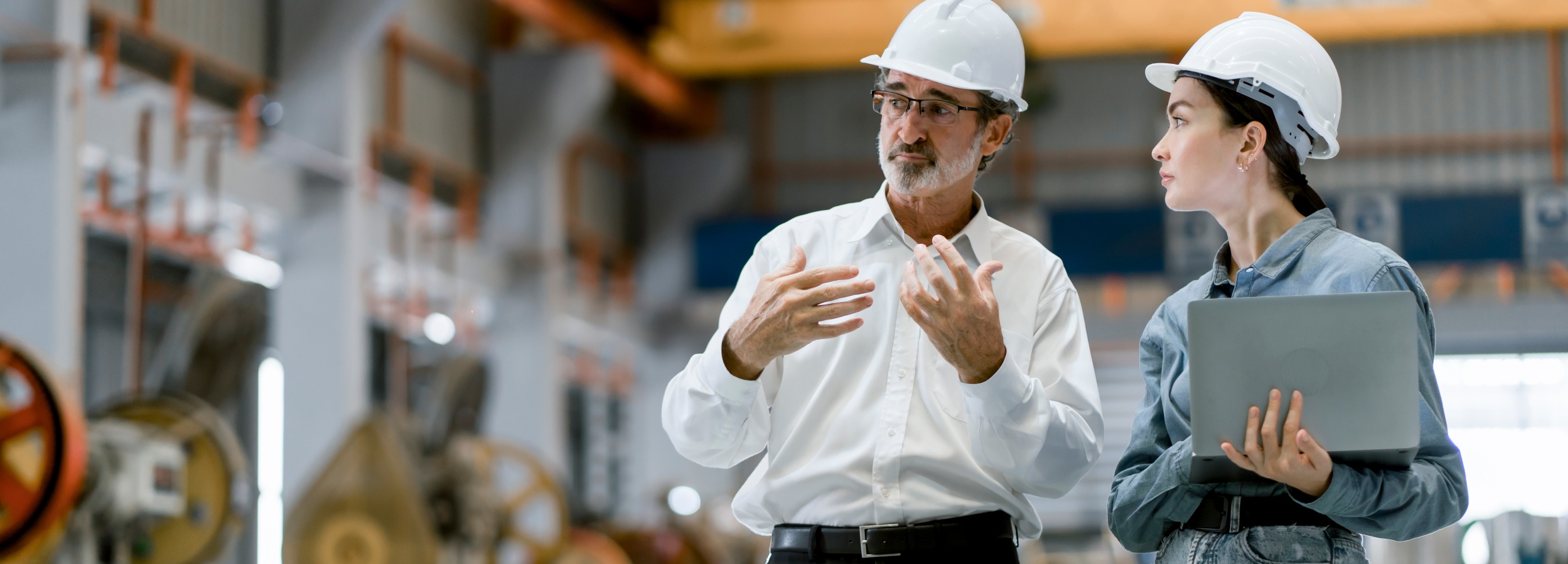 Ein erfahrener Ingenieur und eine jüngere Kollegin besprechen ein Projekt in einer Fabrik.

Das Bild zeigt zwei Personen, die sich in einer Fabrikhalle unterhalten. Im Vordergrund stehen ein älterer Mann und eine junge Frau, beide tragen weiße Schutzhelme. Der Mann, mit Brille und grauem Bart, ist in ein weißes Hemd gekleidet und gestikuliert mit beiden Händen, als würde er etwas erklären. Die Frau, die ihm aufmerksam zuhört, trägt ein blaues Jeanshemd und hält einen grauen Laptop in den Händen. Ihre Kleidung deutet auf eine Arbeitsumgebung hin. Im Hintergrund ist die industrielle Umgebung der Fabrik angedeutet, mit Maschinen und technischen Anlagen, die jedoch unscharf und im Hintergrund gehalten sind, um den Fokus auf die beiden Personen zu lenken. Die Beleuchtung ist neutral und die Atmosphäre wirkt professionell und konzentriert, was auf eine Arbeitsbesprechung oder eine Einweisung hindeutet. Die Stimmung ist ruhig und sachlich.