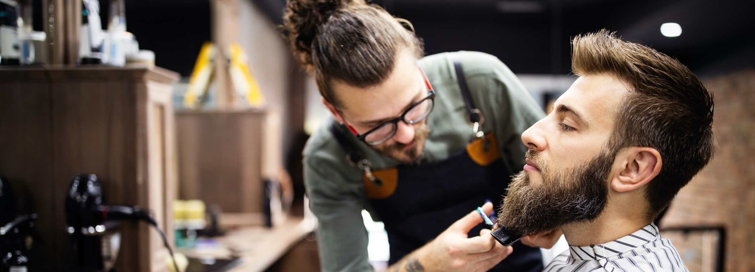Ein Friseur trimmt den Bart eines Kunden in einem Barbershop.

Das Bild zeigt einen Mann mit einem Dutt und Brille, der als Friseur in einem Barbershop tätig ist. Er trägt eine grüne Arbeitskleidung und eine schwarze Schürze. Er steht über einem Kunden und trimmt dessen Bart mit einer Schere und einem Kamm. Der Kunde hat einen vollen, gepflegten Bart und trägt einen Umhang mit schwarzen und weißen Streifen. Die Atmosphäre wirkt professionell und entspannt. Im Hintergrund sind Regale mit Friseurprodukten und Geräten zu sehen, die den Eindruck eines gut ausgestatteten Barbershops verstärken. Die Stimmung ist ruhig und konzentriert, da der Friseur sorgfältig an der Bartpflege arbeitet.