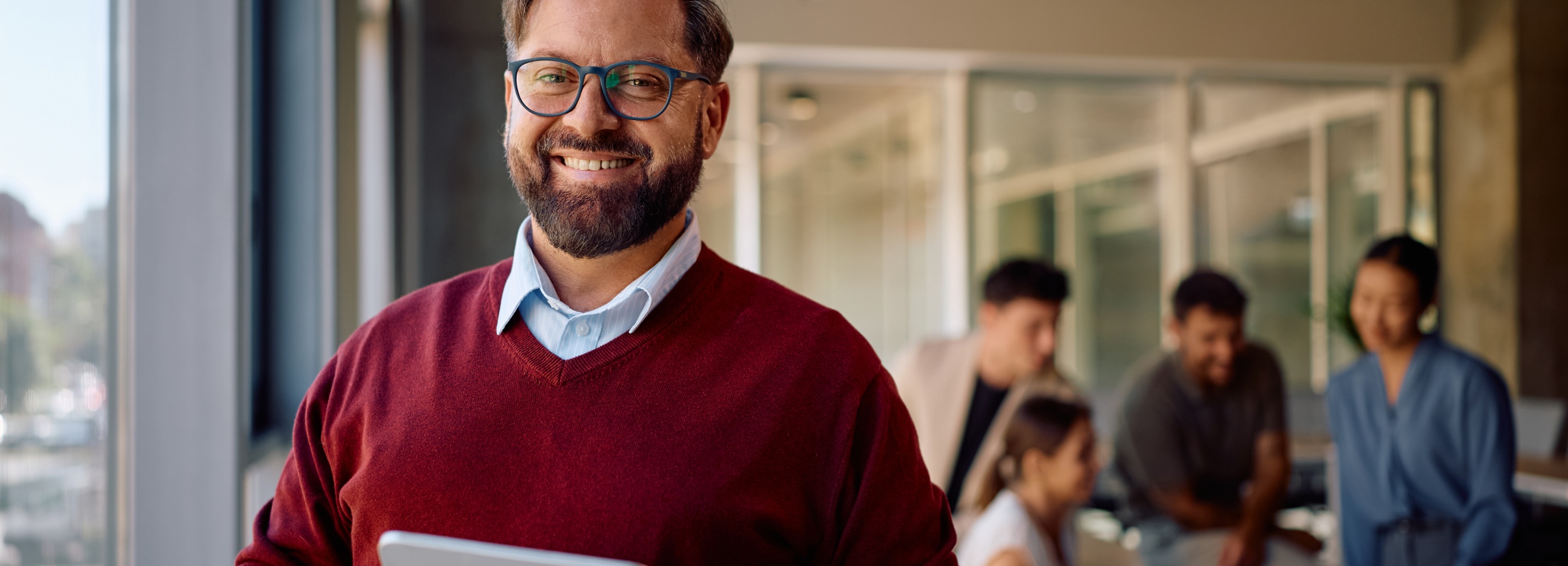 Ein Mann mit Brille lächelt in die Kamera, während er im Hintergrund ein unscharfes Teammeeting leitet.

Das Bild zeigt einen Mann mit Bart und Brille, der freundlich lächelt und ein Tablet hält, während im Hintergrund eine Gruppe von vier Personen in einem Meetingraum zu sehen ist. Der Mann trägt einen roten Pullover über einem hellblauen Hemd. Im Hintergrund sitzen oder stehen die Teammitglieder und scheinen in ein Gespräch vertieft zu sein. Die Atmosphäre wirkt professionell und entspannt zugleich, mit einem Fokus auf Zusammenarbeit und positiver Kommunikation. Das Licht ist hell und natürlich, was die offene und einladende Stimmung des Raumes unterstreicht.