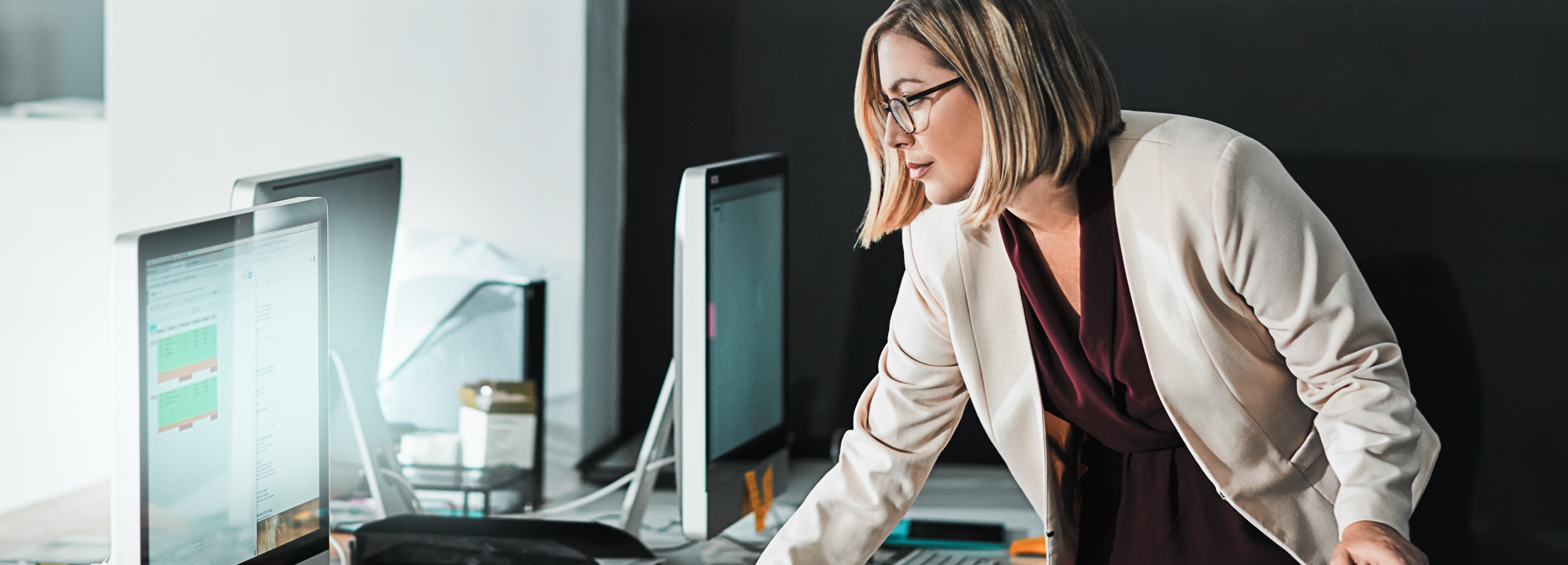 Eine professionell gekleidete Frau beugt sich über einen Schreibtisch voller Computerbildschirme und anderer Bürogegenstände, während sie konzentriert auf einen der Bildschirme schaut.

Das Bild zeigt eine blonde Frau mit Brille, die einen beigen Blazer und eine dunkelrote Bluse trägt. Sie steht an einem Schreibtisch, auf dem mehrere Computerbildschirme stehen. Auf den Bildschirmen sind verschiedene Fenster und Anwendungen zu sehen. Die Frau scheint sich auf den Inhalt eines der Bildschirme zu konzentrieren. Die Atmosphäre des Bildes ist professionell und konzentriert. Der Hintergrund ist unscharf und dunkel, was den Fokus auf die Frau und die Computerbildschirme lenkt. Das Bild vermittelt den Eindruck von harter Arbeit und Engagement im Büro.